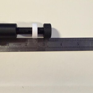 Op de afbeelding: Een zwarte metalen klem met een schroefdraad, een witte ring en een zwarte knop met textuur. Een metalen liniaal met centimeter markeringen ligt naast de klem. De klem is ongeveer 7,6 cm lang.