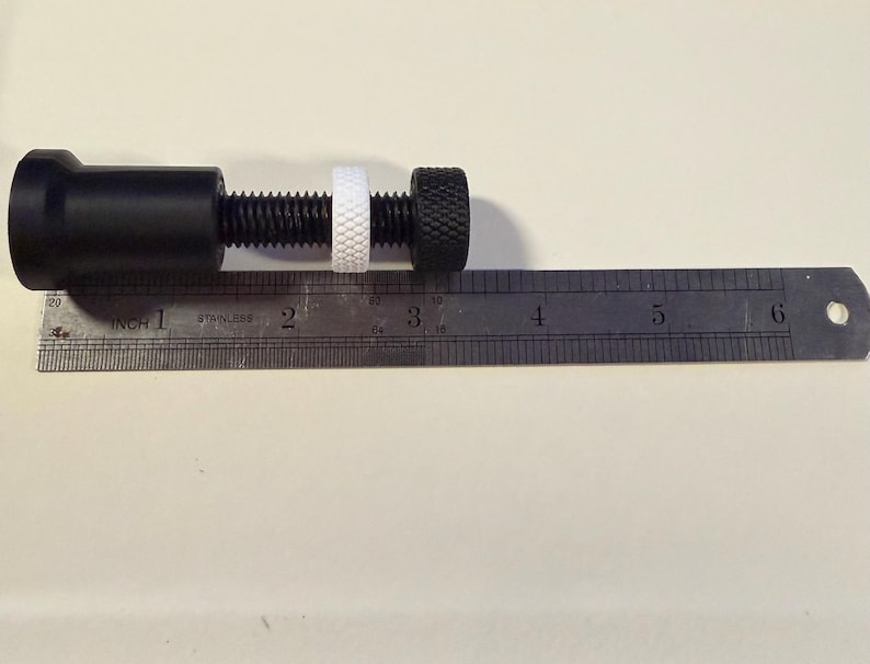 Op de afbeelding: Een zwarte metalen klem met een schroefdraad, een witte ring en een zwarte knop met textuur. Een metalen liniaal met centimeter markeringen ligt naast de klem. De klem is ongeveer 7,6 cm lang.