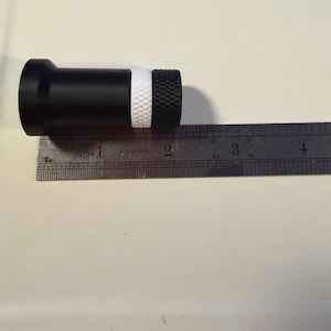 Op de afbeelding: Een zwart cilindrisch object met een witte band en een getextureerd zwart uiteinde, ongeveer 5 cm lang, ligt naast een liniaal. De liniaal is gemarkeerd met inches en het woord "stainless".