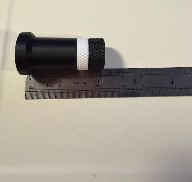 Op de afbeelding: Een zwart cilindrisch object met een witte band en een getextureerd zwart uiteinde, ongeveer 5 cm lang, ligt naast een liniaal. De liniaal is gemarkeerd met inches en het woord "stainless".