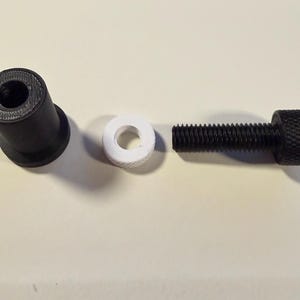 Op de afbeelding: Een gedemonteerde zwarte bout met een gekartelde kop, een witte ring en een zwart cilindrisch onderdeel. De onderdelen zijn gerangschikt op een lichtgekleurd oppervlak.