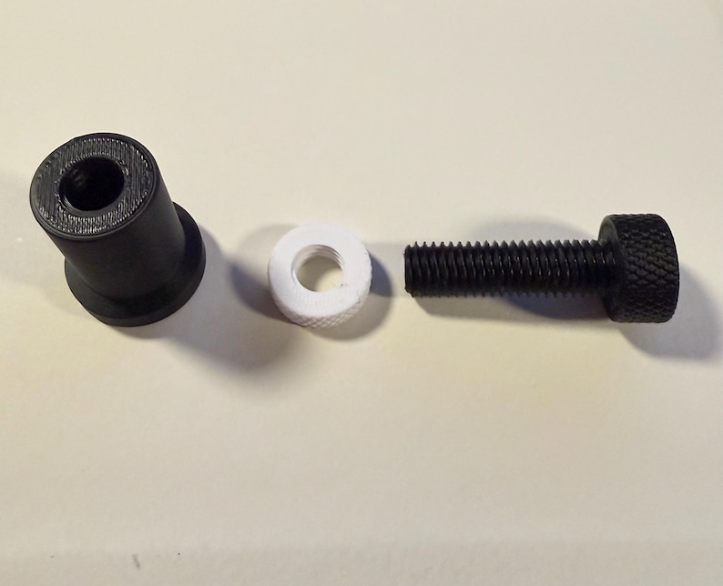 Op de afbeelding: Een gedemonteerde zwarte bout met een gekartelde kop, een witte ring en een zwart cilindrisch onderdeel. De onderdelen zijn gerangschikt op een lichtgekleurd oppervlak.