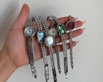 Könnte beinhalten: Eine Sammlung von sechs silberfarbenen Armbanduhren mit verschiedenen Zifferblattformen und -farben. Die Zifferblätter umfassen runde, quadratische und rechteckige Designs mit Farben wie Weiß, Blau, Grün und Burgunderrot. Die Armbänder sind aus Metall gefertigt.