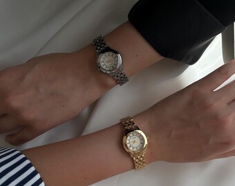 May include: Two wristwatches, one silver and one gold, are displayed. The silver watch has a silver band and a white face with black Roman numerals. The gold watch has a gold band and a white face. Both watches are on wrists.