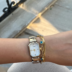 Pode incluir: Um relógio de pulso dourado e prateado com mostrador branco e pulseira dourada. O relógio tem um mostrador retangular com mostrador de madrepérola. Uma pulseira dourada com amuletos de olho grego azuis também é visível.