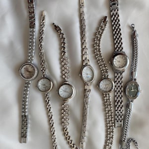 Könnte beinhalten: Sammlung von sieben silberfarbenen Armbanduhren mit verschiedenen Armbanddesigns. Die Zifferblätter der Uhren sind überwiegend weiß oder hell, einige mit dekorativen Elementen. Die Armbänder umfassen Glieder-, Ketten- und verzierte Stile.