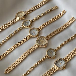 May include: An assortment of gold-toned wristwatches with various band styles and face designs. Some watches feature clear gemstones along the bands. The watch faces are round, oval, and rectangular.
