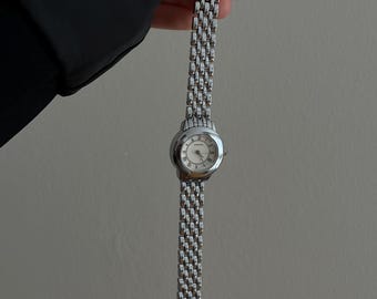 May include: A silver wristwatch with a round face and a linked band. The watch has a light-colored face with Roman numeral hour markers. The band is made of small, rectangular links. The watch is held against a neutral background.