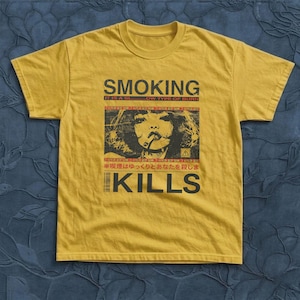 Puede incluir: Una camiseta de manga corta color amarillo mostaza con un estampado gráfico en negro y rojo. El diseño incluye el texto SMOKING KILLS, una ilustración de una persona fumando y caracteres japoneses, con una estética desgastada y vintage.