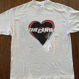 Könnte beinhalten: Weißes T-Shirt mit einem schwarzen Herz-Design mit roter Umrandung. Der Text "THE CURE" ist in Weiß über dem Herz aufgedruckt. Das T-Shirt hat einen Rundhalsausschnitt und kurze Ärmel.