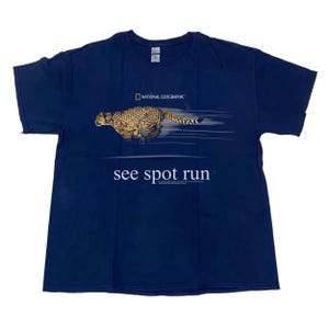 Könnte beinhalten: Ein kurzärmeliges T-Shirt in Marineblau mit einer Grafik eines rennenden Geparden und Bewegungslinien. Über dem Geparden befindet sich das National Geographic Logo. Unter der Grafik steht der Text see spot run in weißen Buchstaben.