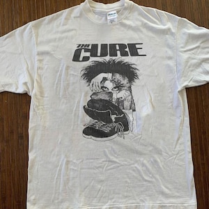 Könnte beinhalten: Weißes T-Shirt mit dem Bandnamen "THE CURE" in Schwarz über einer Schwarz-Weiß-Grafik. Die Grafik zeigt eine Person mit dunklen Haaren und schwarzen Turnschuhen. Das T-Shirt ist aus einem weichen Material.