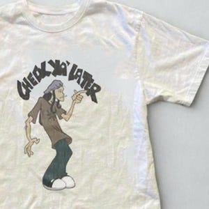 Puede incluir: Una camiseta de manga corta de color crema con una ilustraci&oacute;n gr&aacute;fica de un personaje de dibujos animados se&ntilde;alando con el dedo. Sobre el personaje, el texto "CHECK YA LATER" aparece en una fuente oscura, negrita y desgastada.