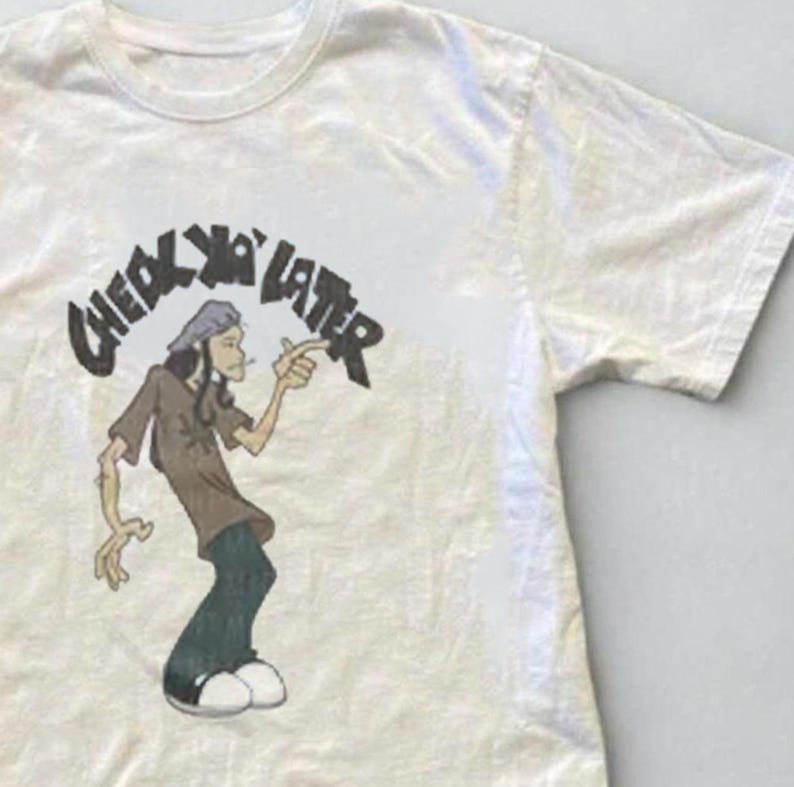 Puede incluir: Una camiseta de manga corta de color crema con una ilustraci&oacute;n gr&aacute;fica de un personaje de dibujos animados se&ntilde;alando con el dedo. Sobre el personaje, el texto "CHECK YA LATER" aparece en una fuente oscura, negrita y desgastada.