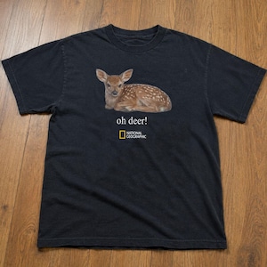 Pode incluir: Uma t-shirt preta de manga curta com um gráfico de um cervo castanho malhado deitado. Abaixo do cervo, o texto "oh deer!" aparece em branco, seguido pelo logótipo da National Geographic em amarelo e branco centrado no peito.