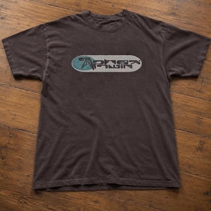 Könnte beinhalten: Ein dunkelbraunes Kurzarm-T-Shirt mit einer Grafik auf der Brust. Die Grafik besteht aus einem hellblauen Kreis, der einen weißen, horizontalen, pillenförmigen Hintergrund überlagert, mit dem schwarzen Text Aphex Twin in der Mitte.