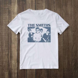 Könnte beinhalten: Ein weißes kurzärmeliges T-Shirt mit einem blauen Grafikdruck auf der Brust. Die Grafik zeigt den Schriftzug THE SMITHS über einem verwaschenen, einfarbigen Foto der vier Bandmitglieder, wobei der Sänger in der Mitte positioniert ist.