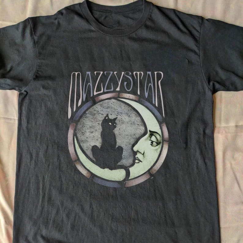 May include: Dark grey t-shirt with the band name "MAZZYSTAR" above a circular graphic. The graphic shows a black cat on a crescent moon with a face, set against a grey and blue background.