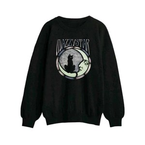 Pode incluir: Camisola preta com o texto "MAZZYSTAR" acima de um gráfico de um gato preto sentado em uma lua crescente. A lua tem um rosto e é cercada por uma borda colorida.