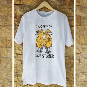 Könnte beinhalten: Weißes T-Shirt mit einer humorvollen Grafik von zwei gelben Vögeln mit schwarzen Umrissen und dem Text "TWO BIRDS ONE STONED" in Schwarz. Die Vögel haben ein cartoonartiges Design.