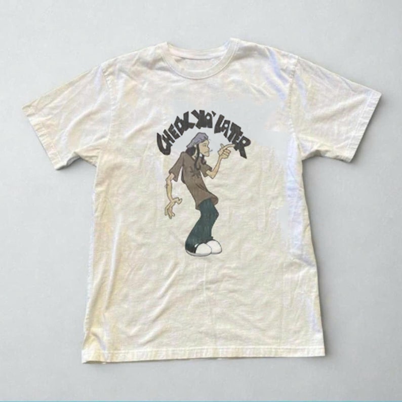 Puede incluir: Una camiseta de manga corta color crema con una ilustraci&oacute;n gr&aacute;fica de un personaje de dibujos animados con camisa marr&oacute;n, pantalones oscuros y sombrero gris. El texto "CHECK YA LATER" aparece en una fuente negra y arqueada arriba.