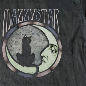 May include: Dark grey t-shirt with the band name MAZZYSTAR above a circular graphic. The graphic shows a black cat on a crescent moon with a face, within a stained-glass style frame. A music-themed item.