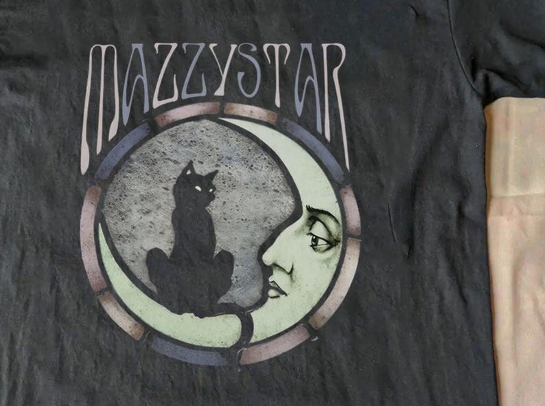 May include: Dark grey t-shirt with the band name MAZZYSTAR above a circular graphic. The graphic shows a black cat on a crescent moon with a face, within a stained-glass style frame. A music-themed item.
