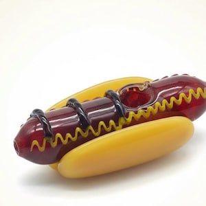 Peut inclure: Une pipe en verre fantaisie en forme de hot-dog. La pipe présente une « saucisse » en verre rouge avec des lignes en zigzag jaunes et des spirales noires, nichée dans un « pain » en verre jaune. Le design est fantaisiste et détaillé.