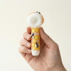 May include: A white and yellow glass pipe with a honeycomb design and three small bee decorations. The pipe is held in a hand against a plain background. The honeycomb pattern is yellow with orange outlines.