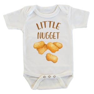 Op de afbeelding: Witte babybody met de woorden "LITTLE NUGGET" in bruine letters. De body heeft een aquarel illustratie van verschillende goudbruine kipnuggets. De body heeft een drukknoopsluiting aan de onderkant.