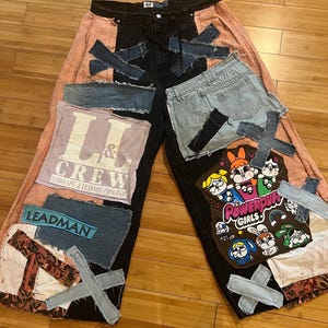 May include: Custom patchwork jeans with a black center panel, peach side panels, and various denim patches. Features patches with text and cartoon characters. The jeans have a wide-leg design.