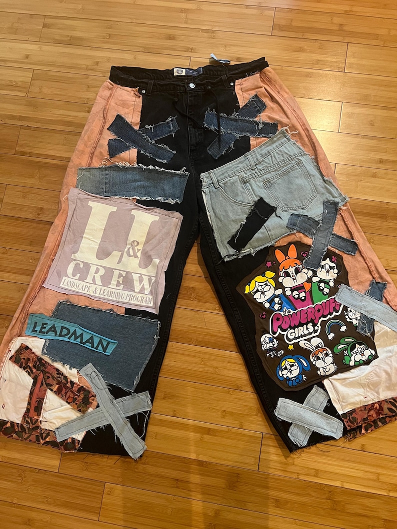 May include: Custom patchwork jeans with a black center panel, peach side panels, and various denim patches. Features patches with text and cartoon characters. The jeans have a wide-leg design.