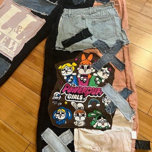 May include: A patchwork design featuring denim shorts, a pink t-shirt with text, and a brown patch with Powerpuff Girls characters. Denim patches are sewn onto black and pink fabric.