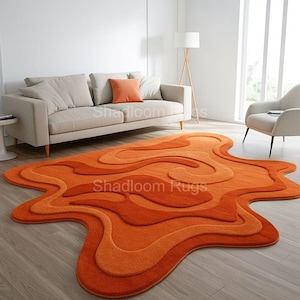May include: A vibrant orange and rust-toned rug with a distinctive, flowing design. The rug showcases layered, swirling patterns in various shades of orange, creating a three-dimensional effect. The rug is placed on a wooden floor in a living room.