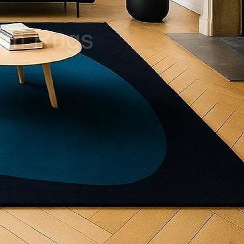 May include: A modern area rug with a dark navy blue background and a large, teal blue oval design. The rug is placed on a hardwood floor, with a round wooden coffee table and a black decorative object in the background.