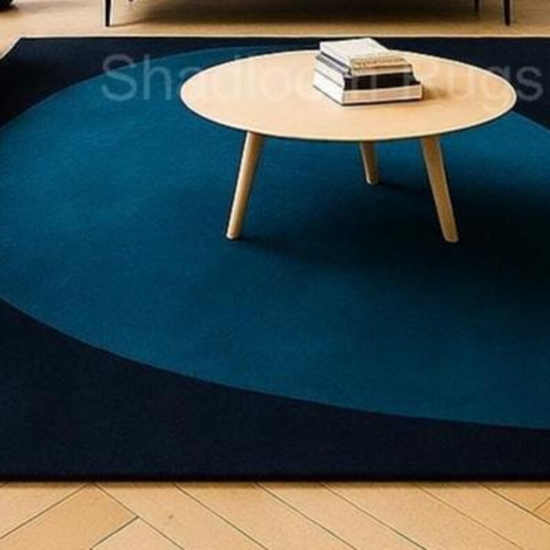 May include: A modern area rug with a dark blue and navy blue color scheme. A light wood-toned round coffee table with three legs sits on the rug. Several books are stacked on the table. The rug has a unique geometric design.