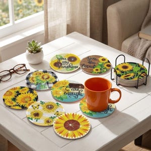 Op de afbeelding: Een set ronde diamond painting onderzetters met zonnebloemmotieven en teksten zoals you are my sunshine, you are loved en you are enough. De set bevat een zwarte metalen houder en een oranje mok die op een van de onderzetters staat.