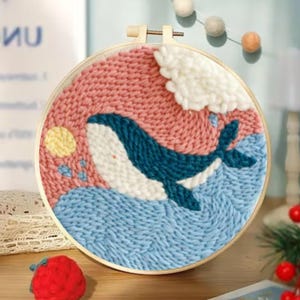 May include: A handcrafted embroidery hoop art piece featuring a whale design. The whale is depicted in shades of blue and white, swimming in blue waves under a pink sky with a yellow sun and white cloud. The artwork is set in a wooden hoop.