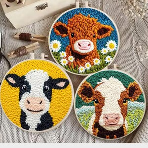 May include: Three embroidered cow portraits in round hoops. One features a brown cow with daisies on a blue background, another a black and white cow on yellow, and the third a brown and white cow on green.