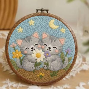 May include: Embroidered artwork featuring two gray kittens holding flowers, set against a blue sky with stars and a crescent moon. The kittens have pink ears and are smiling. The artwork is framed in a wooden hoop.