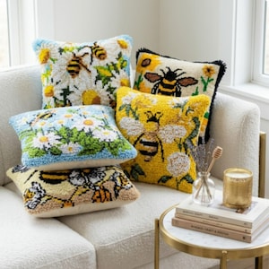 May include: A collection of four square throw pillows featuring textured punch needle designs of bees and flowers in yellow, white, blue, and green. The pillows are arranged on a sofa, showcasing various patterns of insects and floral motifs.