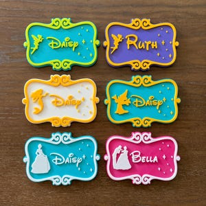 May include: Six colorful name tags with Disney-themed designs. Each tag features a different color scheme, including teal, purple, white, and pink, with names like Daisy, Ruth, and Bella. The tags have decorative borders and character illustrations.