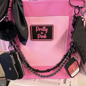May include: A bright pink tote bag featuring a rectangular black patch with the text Pretty in Pink. The bag is decorated with black accessories including a fluffy pom-pom, a satin ribbon, a small zippered pouch, a chain, and a hand sanitizer holder.