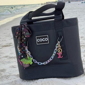 May include: A black tote bag featuring a rectangular patch with the text "coco" in white. The bag is adorned with a black patterned ribbon, a bright green alligator charm, a pastel-colored plastic chain, and a pink logo charm attached to the side.