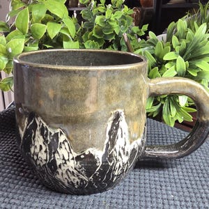 May include: A ceramic mug featuring a two-tone glaze with a glossy olive green top and a dark charcoal base. The lower section is decorated with a carved, white-highlighted mountain range design that wraps around the exterior of the vessel.