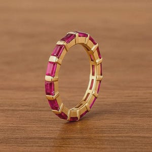 May include: A gold ring featuring a continuous band of rectangular, magenta gemstones. The ring's design showcases a repeating pattern of the gemstones set within a gold band, creating a vibrant and eye-catching piece of jewellery.