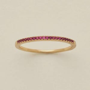 May include: A gold band ring set with small, vibrant pink gemstones. The ring features a delicate design with the gemstones set along the top of the band. The ring is shown against a neutral background.