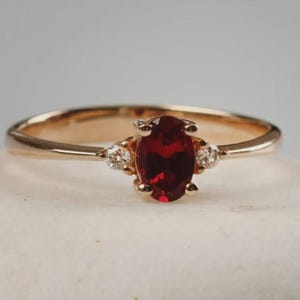 May include: A gold ring featuring an oval-cut, deep red gemstone as the centerpiece, flanked by two smaller, round, clear gemstones. The ring's band is a simple, polished gold. The setting highlights the vibrant red of the main stone.