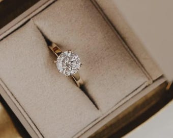 May include: A gold ring with a large, round, clear diamond set in a four-prong setting. The ring is displayed in a beige ring box with a soft, velvety interior.