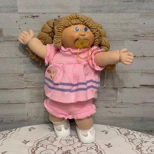 May include: A doll with blonde yarn hair styled in pigtails, blue eyes, and a tan complexion. The doll wears a pink knitted top with a small yellow duck graphic and two purple horizontal stripes, matching pink shorts, and white shoes with a pacifier.